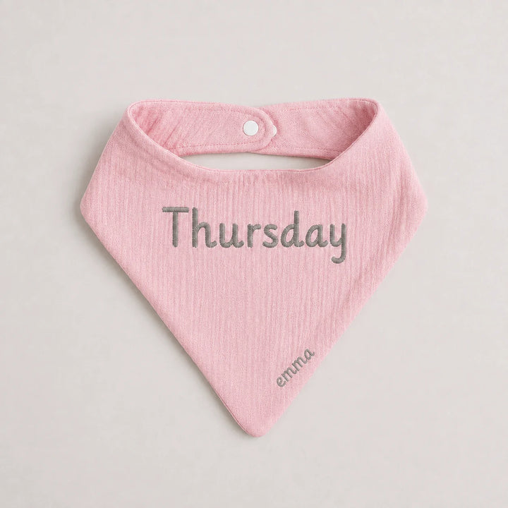 A light pink textured baby bandana bib with 'Thursday' embroidered in gray across the center and 'emma' in a smaller font near the bottom tip, shown on a neutral light gray background.