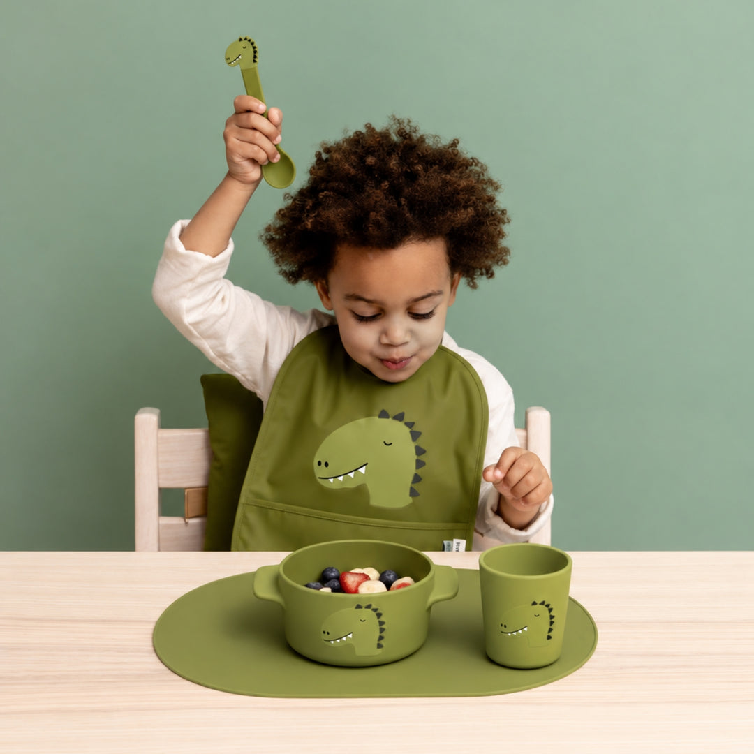 Young child wearing a green dinosaur bib and using the matching Trixie dinosaur bowl, cup and spoon at a wooden table.