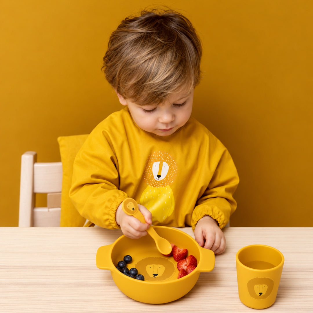 Young child using a mustard yellow Trixie lion mealtime set with matching bowl, cup and spoon at a wooden table.