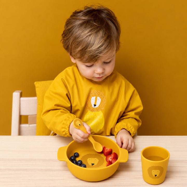Young child using a mustard yellow Trixie lion mealtime set with matching bowl, cup and spoon at a wooden table.