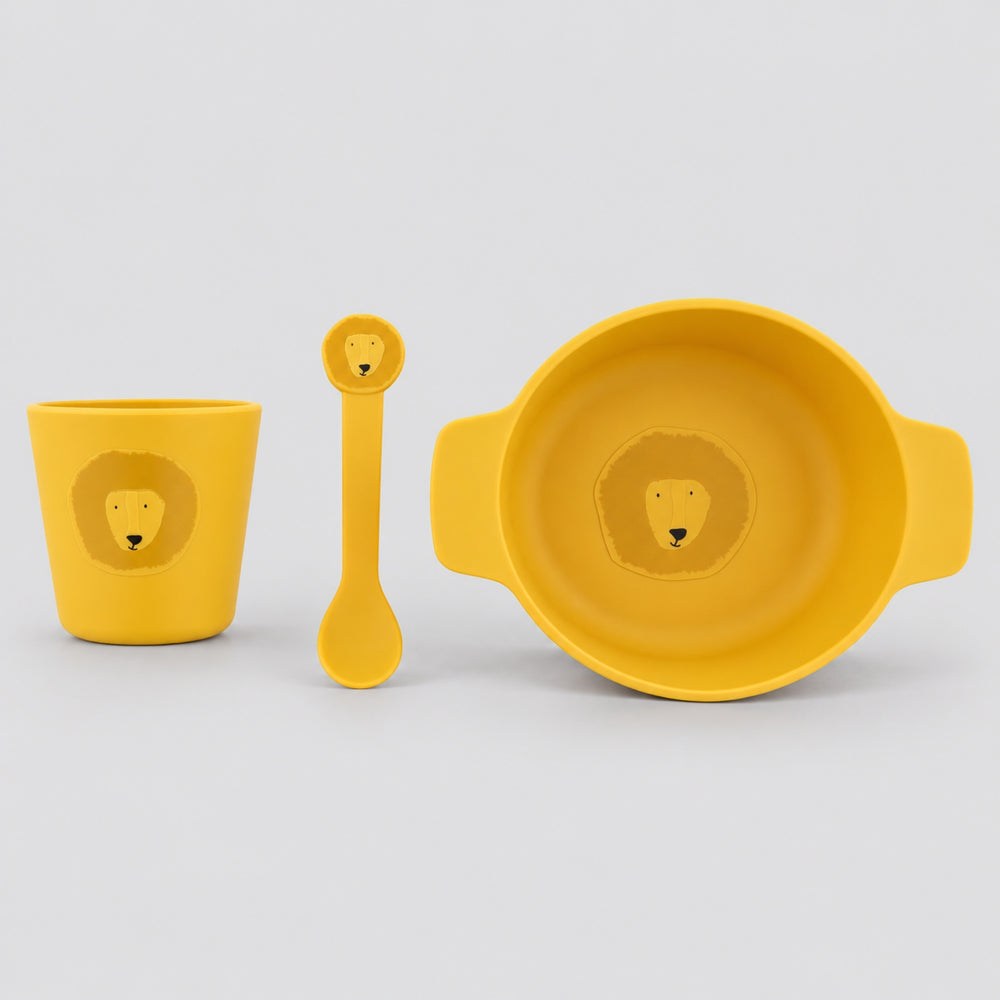 Mustard yellow Trixie lion silicone mealtime set with handled bowl, cup and matching spoon on a light grey studio background.