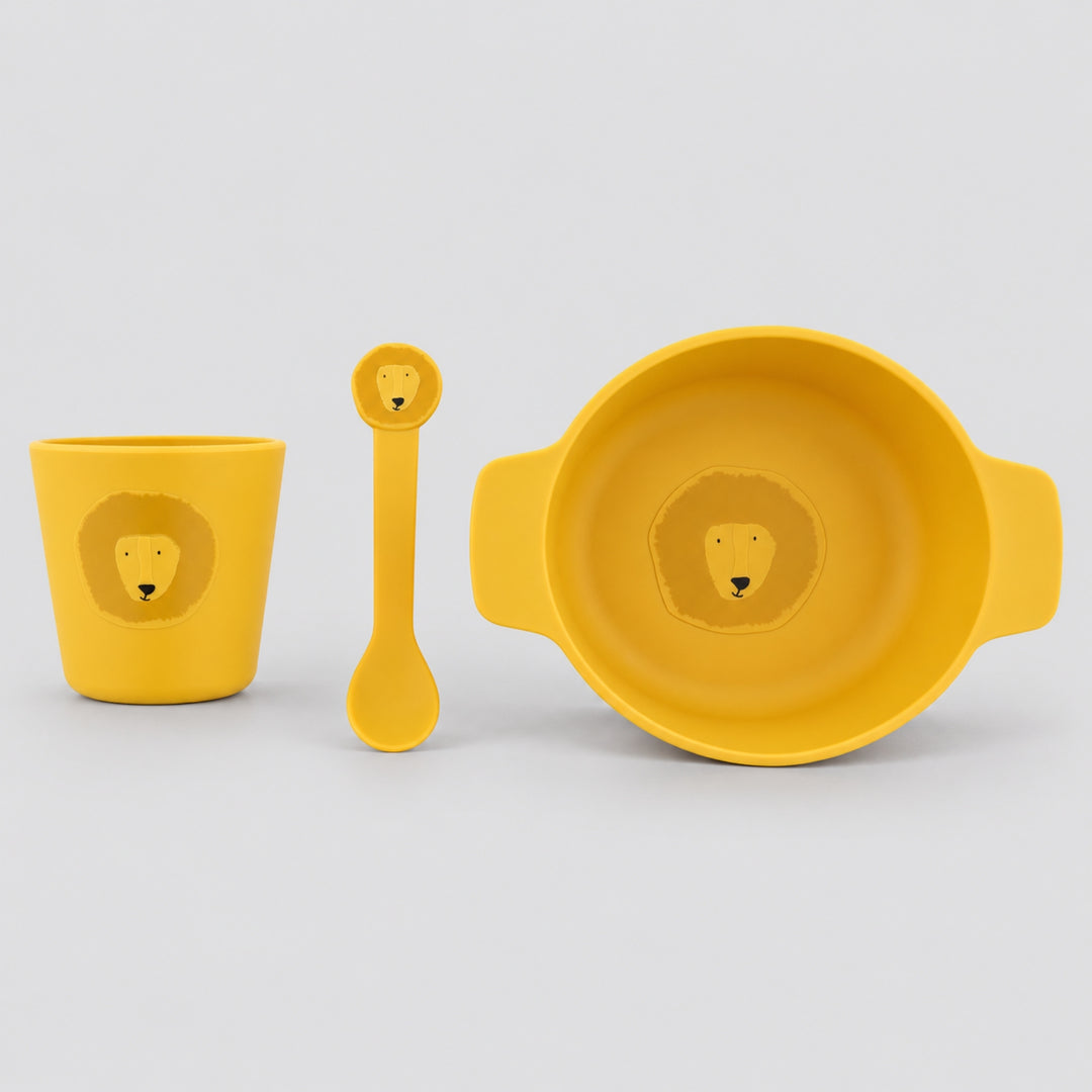 Mustard yellow Trixie lion silicone mealtime set with handled bowl, cup and matching spoon on a light grey studio background.