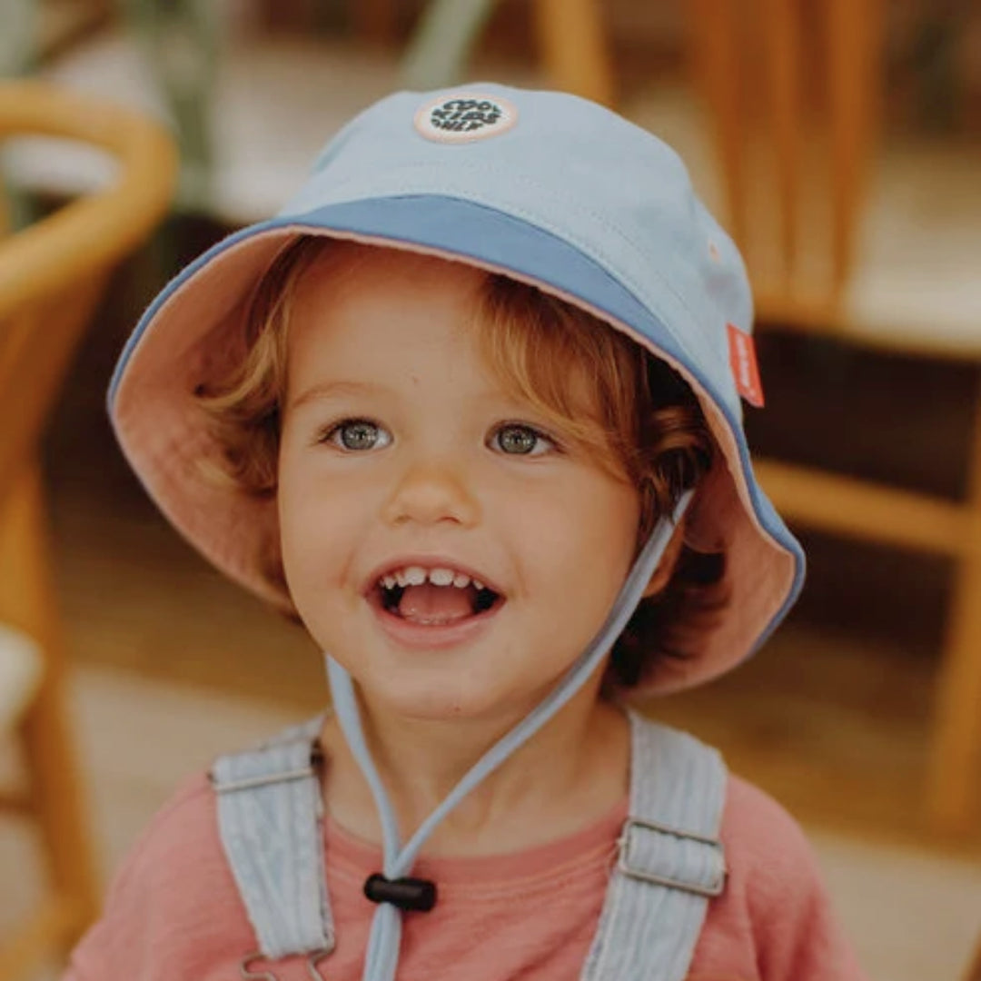 Young child wearing a blue Hello Hossy bucket hat with chin strap in a warm indoor setting.