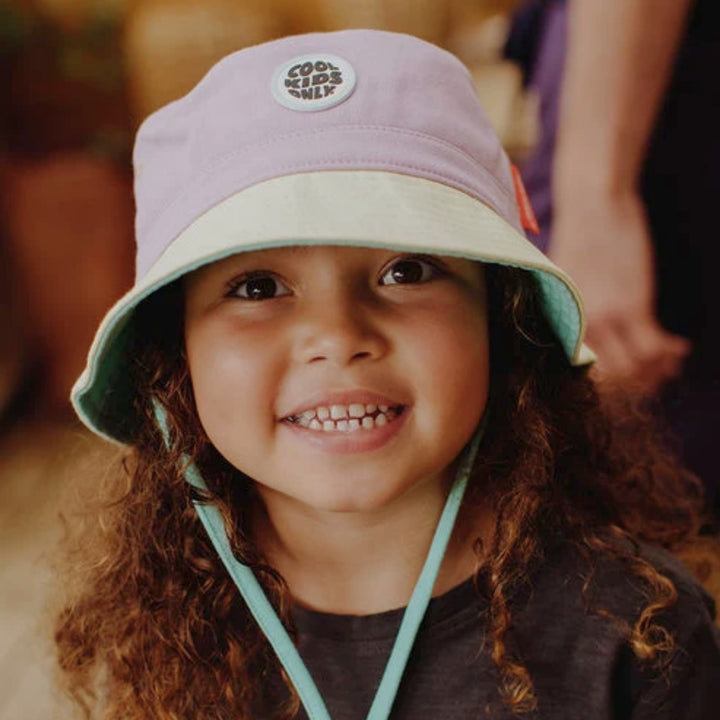 Smiling child wearing a lilac and cream Hello Hossy bucket hat with mint chin strap.
