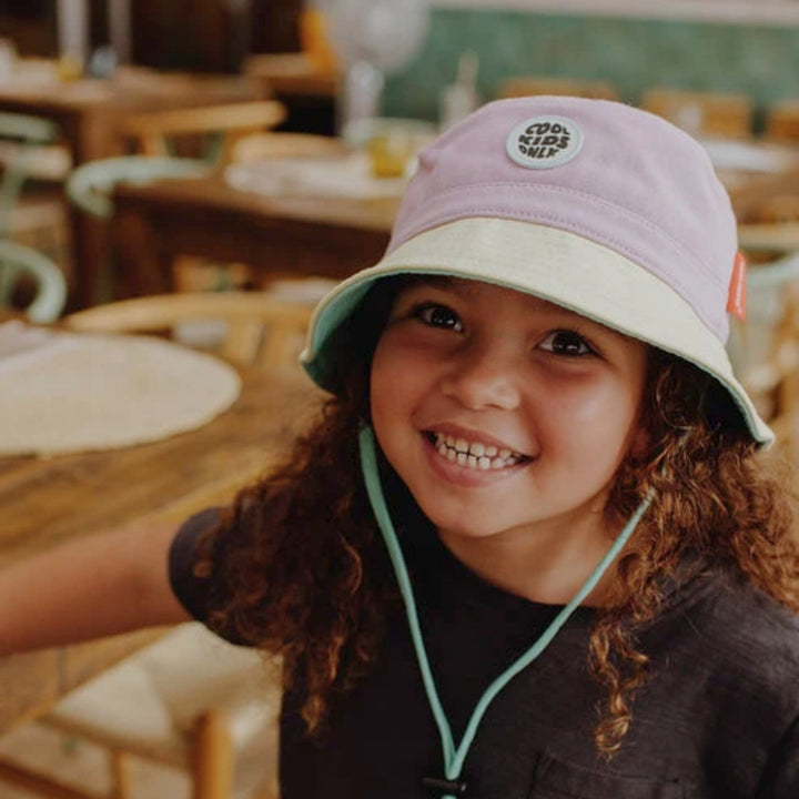 Smiling child wearing a lilac and cream Hello Hossy bucket hat in a warm indoor setting.