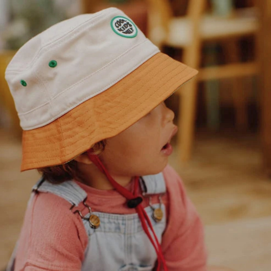 Young child wearing the cream and brown Hello Hossy bucket hat in a warm indoor setting.