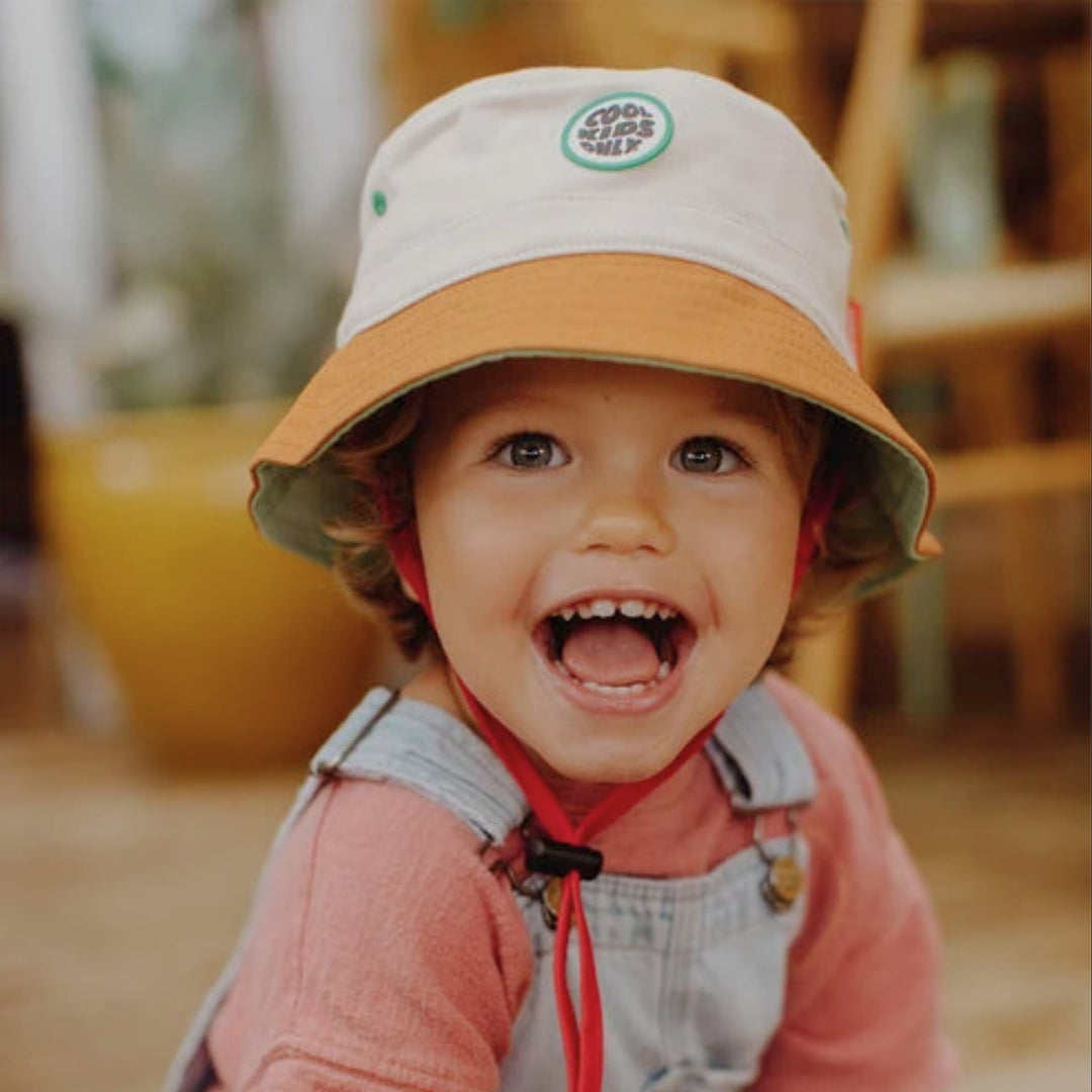 Smiling toddler wearing the cream and brown Hello Hossy bucket hat with red chin strap.