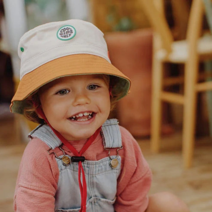 Smiling toddler wearing a cream and brown Hello Hossy bucket hat with red chin strap.