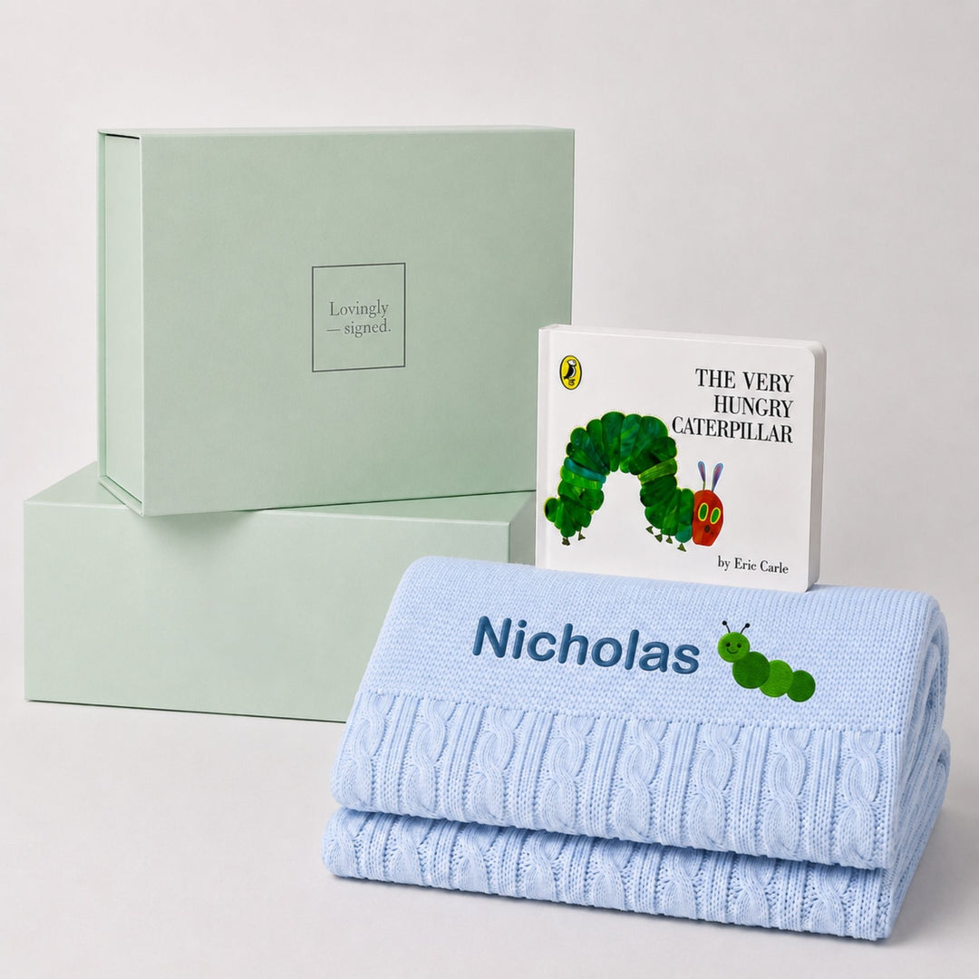Personalised blue cable knit baby blanket embroidered with the name Nicholas and a caterpillar motif, paired with The Very Hungry Caterpillar and a Lovingly Signed gift box.