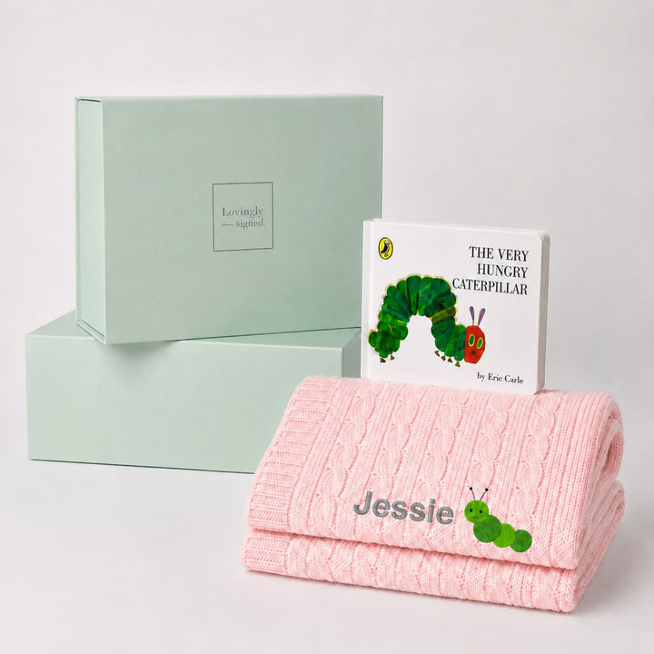 Personalised pink cable knit baby blanket embroidered with the name Jessie and a caterpillar motif, paired with The Very Hungry Caterpillar and a Lovingly Signed gift box.