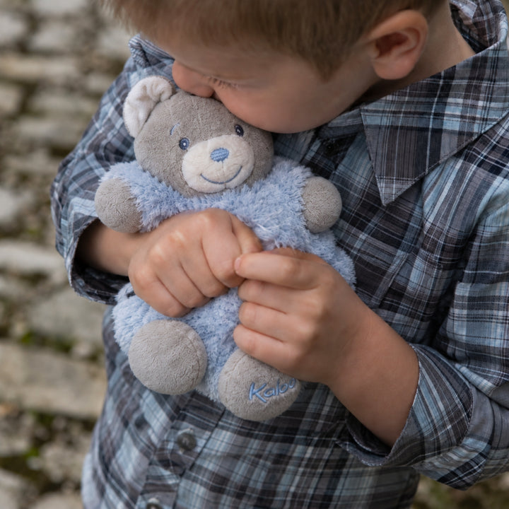 Young boy cuddling a small blue and grey Kaloo teddy bear outdoors.