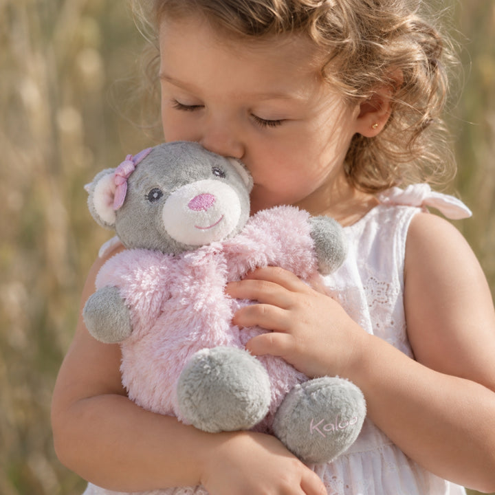 Young girl cuddling a small pink and grey Kaloo teddy bear outdoors.