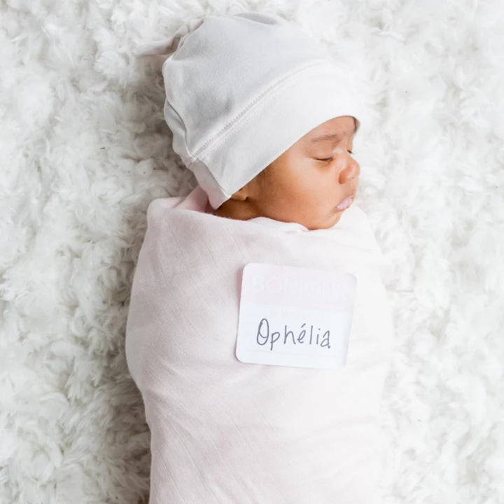 Newborn baby wrapped in a soft pink swaddle and wearing a matching hat, resting on a fluffy white blanket.