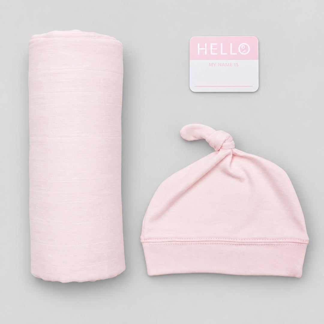 Pink Lulujo Hello World newborn set with rolled muslin swaddle, matching knotted hat and “Hello, my name is” announcement card on a light grey background.