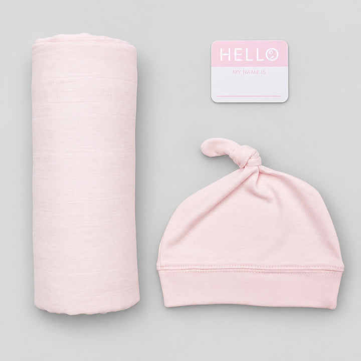 Pink Lulujo Hello World newborn set with rolled muslin swaddle, matching knotted hat and “Hello, my name is” announcement card on a light grey background.
