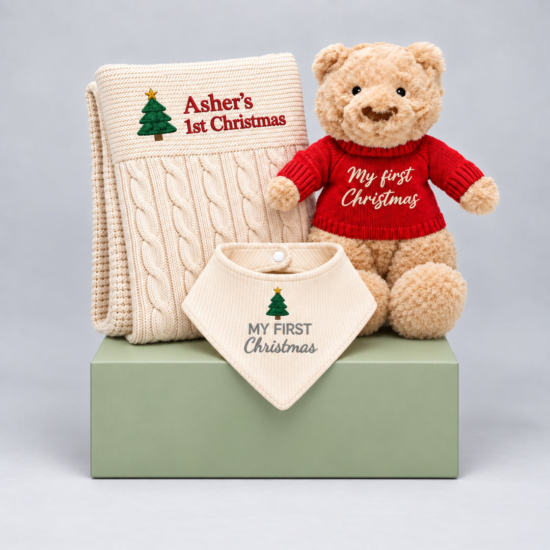 1. Christmas baby gift set featuring a cream cable knit blanket embroidered with a Christmas tree and “Asher’s 1st Christmas,” a teddy bear wearing a red “My First Christmas” jumper, and a matching cream “My First Christmas” bib, arranged on a sage green gift box.
