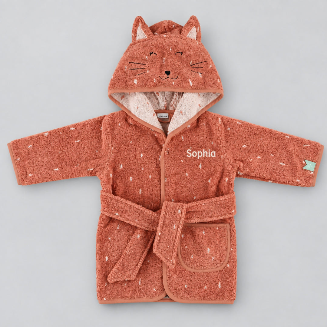 Front view of a terracotta pink children’s cat bathrobe laid flat on a seamless light grey studio background. The robe features a cat-face hood with embroidered facial details and ears, white speckled pattern, matching belt and pocket, personalised with the name “Sophia” in cream embroidery on the chest.