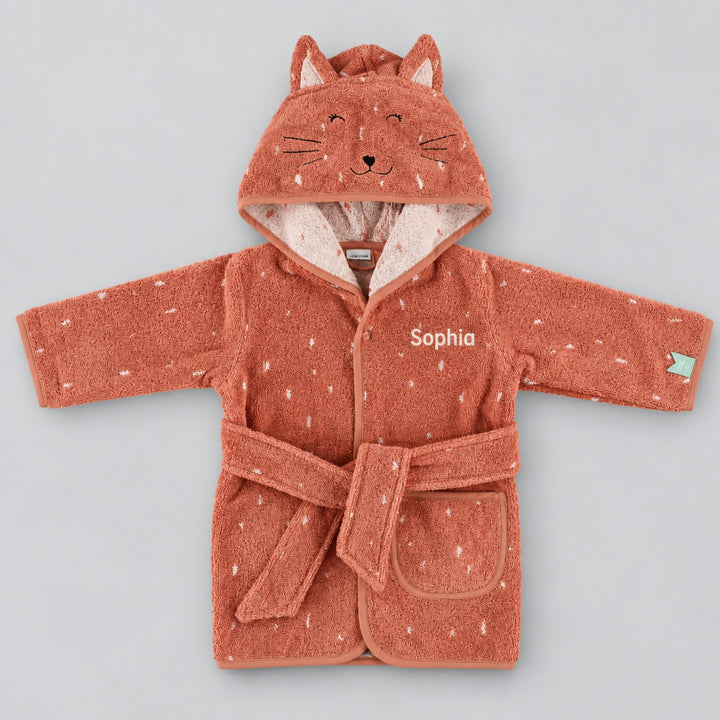 Front view of a terracotta pink children’s cat bathrobe laid flat on a seamless light grey studio background. The robe features a cat-face hood with embroidered facial details and ears, white speckled pattern, matching belt and pocket, personalised with the name “Sophia” in cream embroidery on the chest.