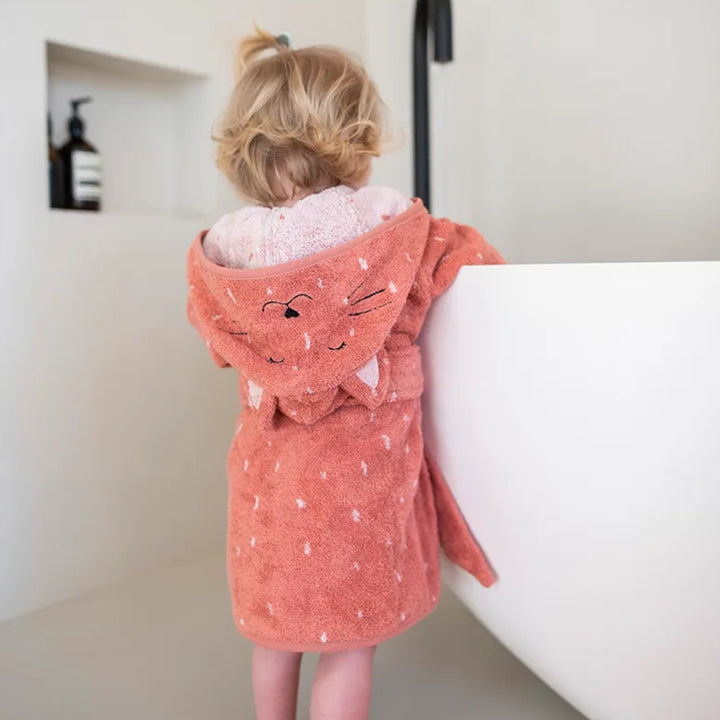 A young child photographed from behind wearing a terracotta pink cat bathrobe beside a white bathtub. The soft terry robe features a playful cat-face hood with embroidered whiskers and ears, a white speckled pattern, and a matching waist belt.