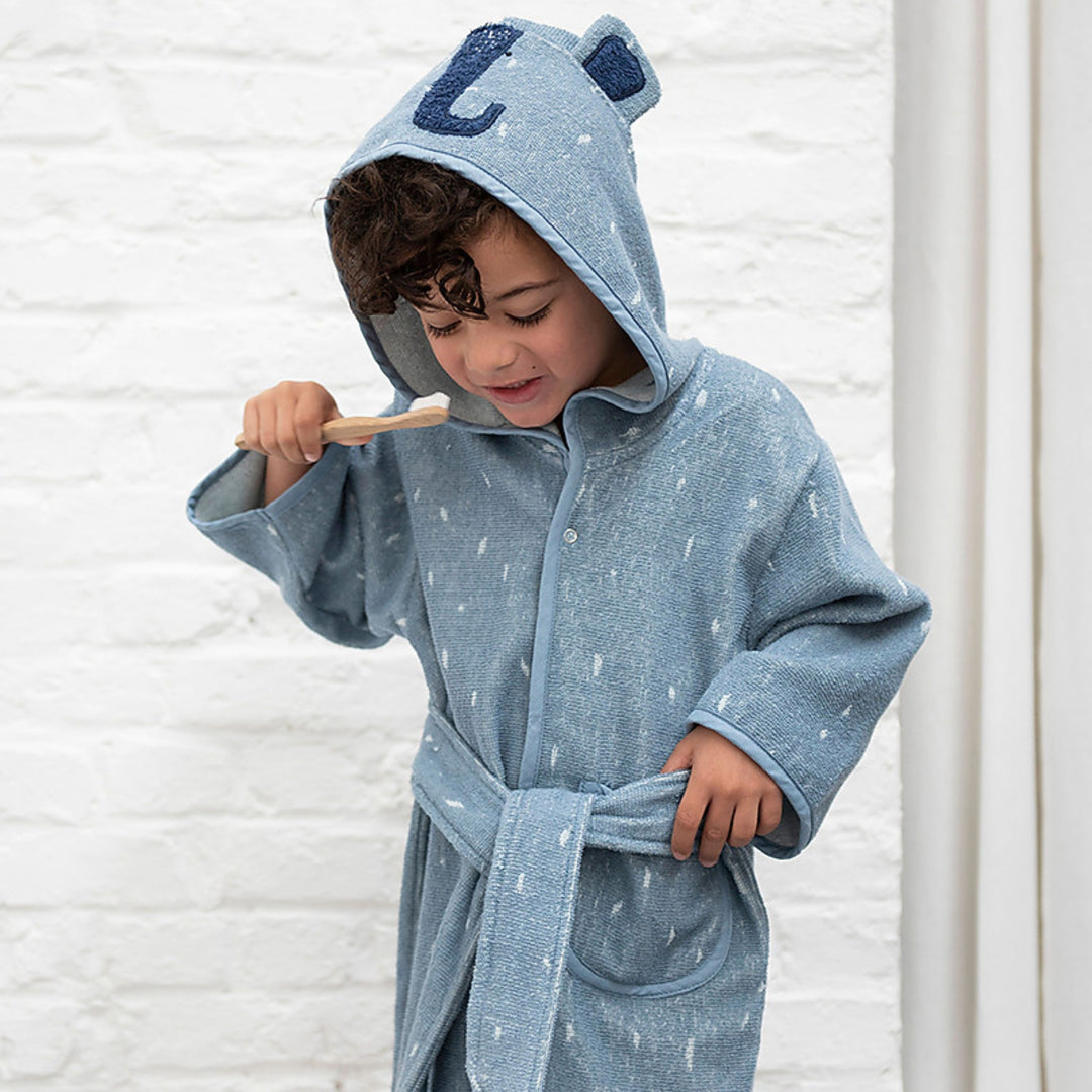 A young boy wearing the dusty blue elephant bathrobe while brushing his teeth. The robe has a hood with rounded elephant ears and a navy trunk detail, white speckled pattern, matching belt, and pocket. The image is photographed in a bright, clean bathroom setting.
