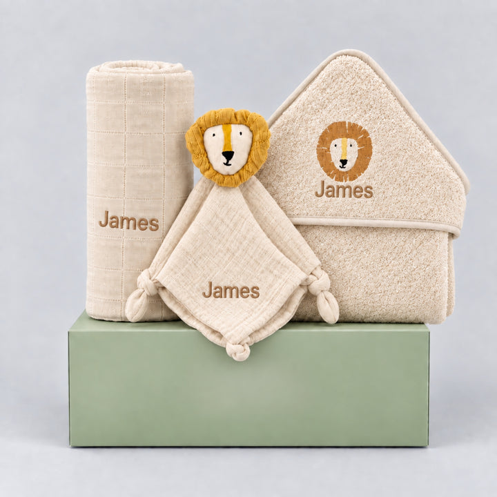Personalised neutral baby gift hamper featuring a rolled cream blanket, a lion comforter and a matching lion hooded towel, all personalised with the name “James” in beige embroidery, arranged on a sage green gift box against a light grey studio background.