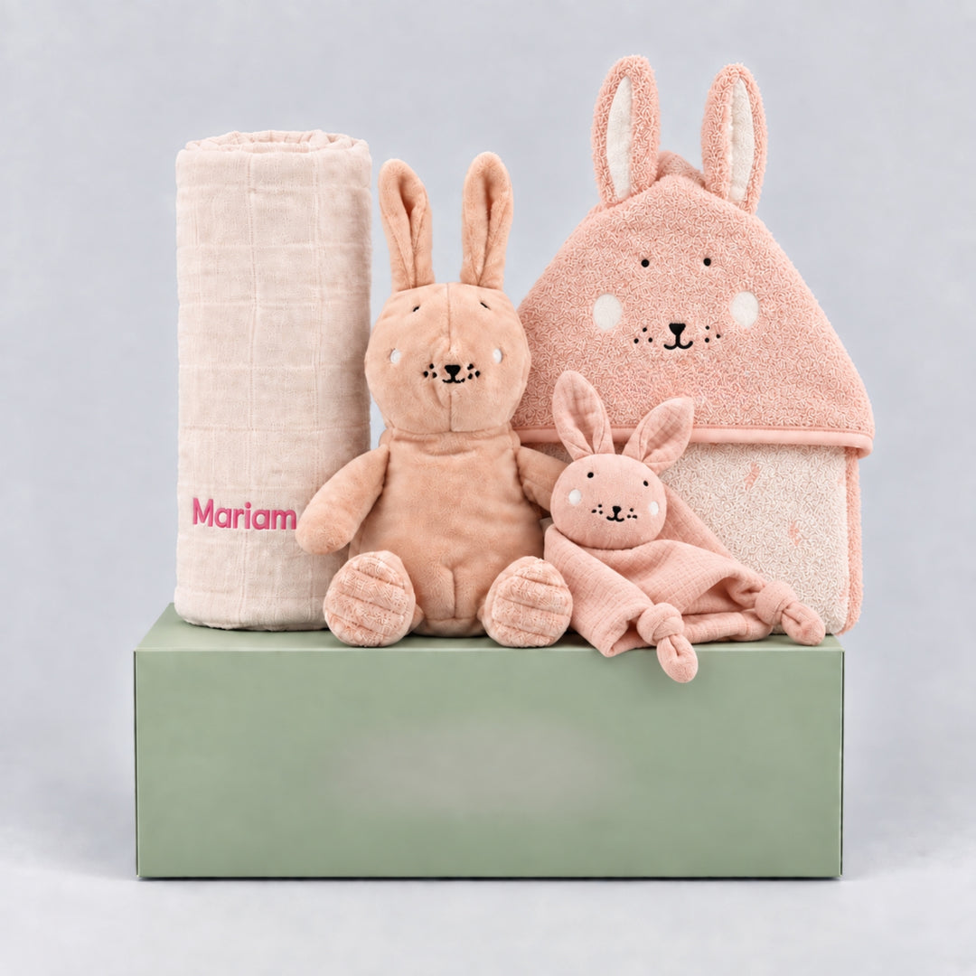 Personalised baby girl gift hamper featuring a rolled cream blanket embroidered with “Mariam” in pink, a soft blush bunny toy, a pink bunny comforter and a matching bunny hooded towel, arranged on a sage green gift box against a light grey studio background.