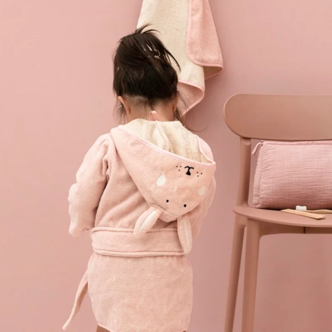 A young child photographed from behind wearing the blush-pink rabbit bathrobe in a coordinated pink bathroom setting. The hood is down across the child’s back, showing the embroidered rabbit face and long bunny ears, with the matching waist belt tied around the robe.