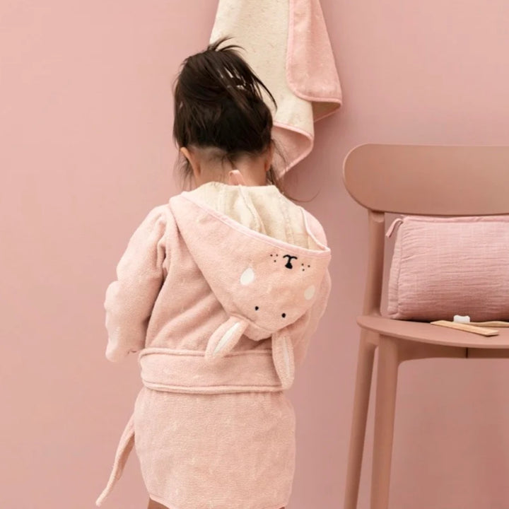 A young child photographed from behind wearing the blush-pink rabbit bathrobe in a coordinated pink bathroom setting. The hood is down across the child’s back, showing the embroidered rabbit face and long bunny ears, with the matching waist belt tied around the robe.