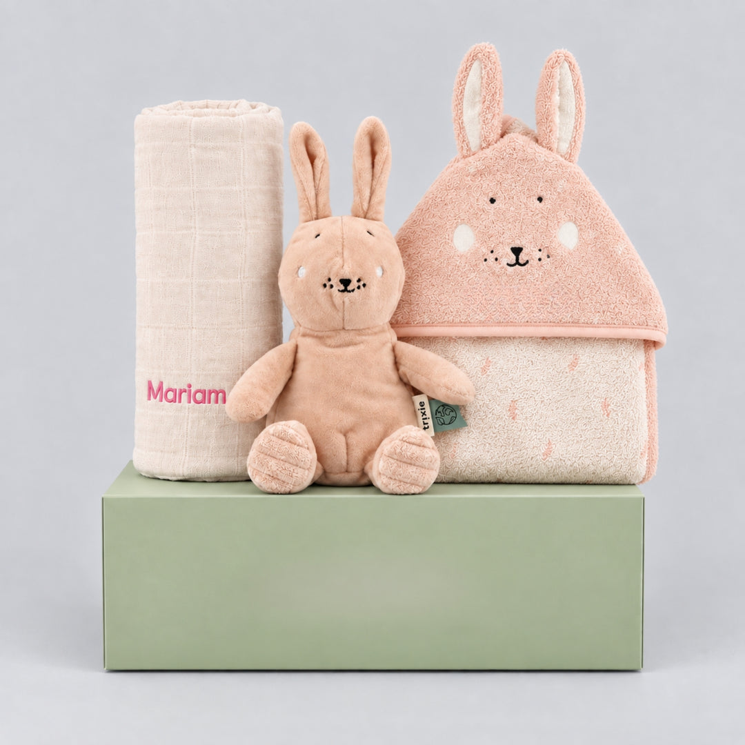 Personalised Trixie bunny gift hamper for a baby girl, featuring a rolled cream blanket embroidered with “Mariam” in pink, a soft blush bunny toy, and a pink bunny hooded towel, arranged on a sage green gift box against a light grey studio background.