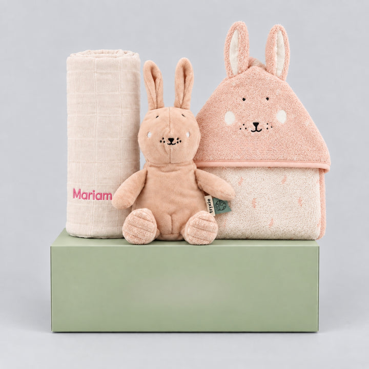 Personalised Trixie bunny gift hamper for a baby girl, featuring a rolled cream blanket embroidered with “Mariam” in pink, a soft blush bunny toy, and a pink bunny hooded towel, arranged on a sage green gift box against a light grey studio background.