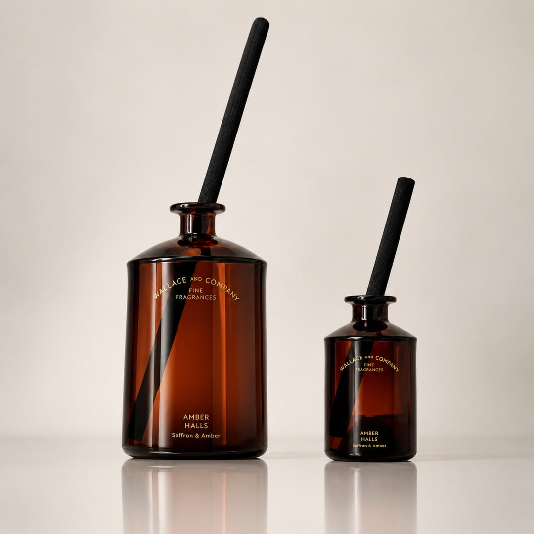 Large and small Wallace and Company “Amber Halls – Saffron & Amber” reed diffusers in dark amber glass bottles with black diffuser sticks, displayed side by side against a warm neutral studio background.