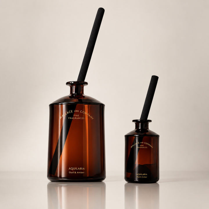 Large and small Wallace and Company “Aquilaria – Oud & Amber” reed diffusers in dark amber glass bottles with black diffuser sticks, displayed side by side against a warm neutral studio background.