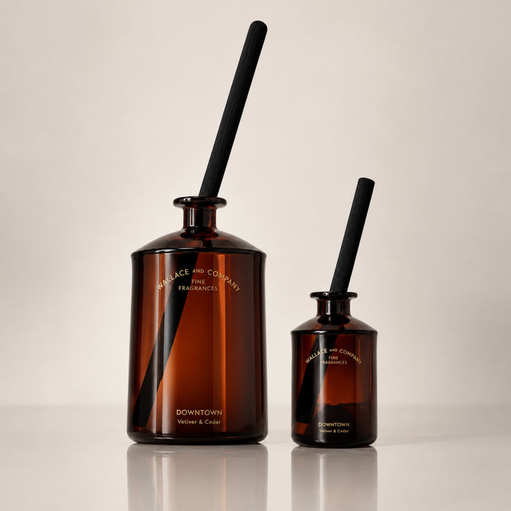 Large and small Wallace and Company “Downtown – Vetiver & Cedar” reed diffusers in amber-brown glass bottles with black diffuser sticks, displayed side by side against a warm neutral studio background.