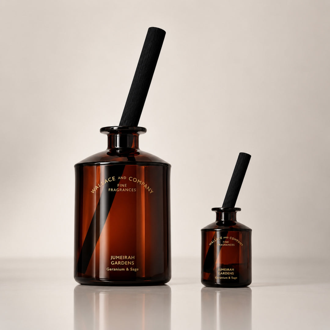 Large and small Wallace and Company “Jumeirah Gardens – Geranium & Sage” reed diffusers in dark amber glass bottles with black diffuser sticks, displayed side by side against a warm neutral studio background.