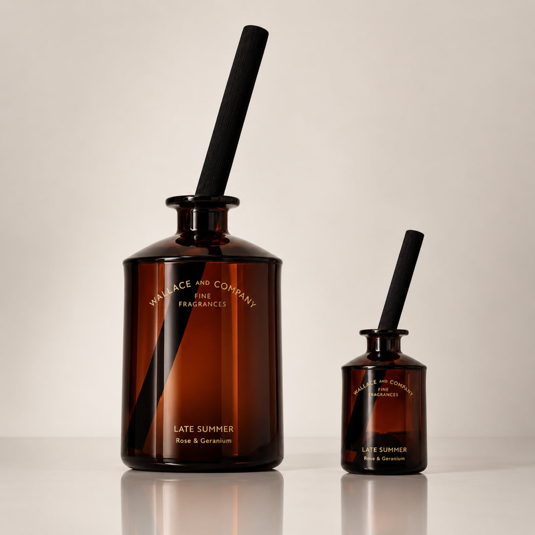 Large and small Wallace and Company “Late Summer – Rose & Geranium” reed diffusers in dark amber glass bottles with black diffuser sticks, displayed side by side against a warm neutral studio background.