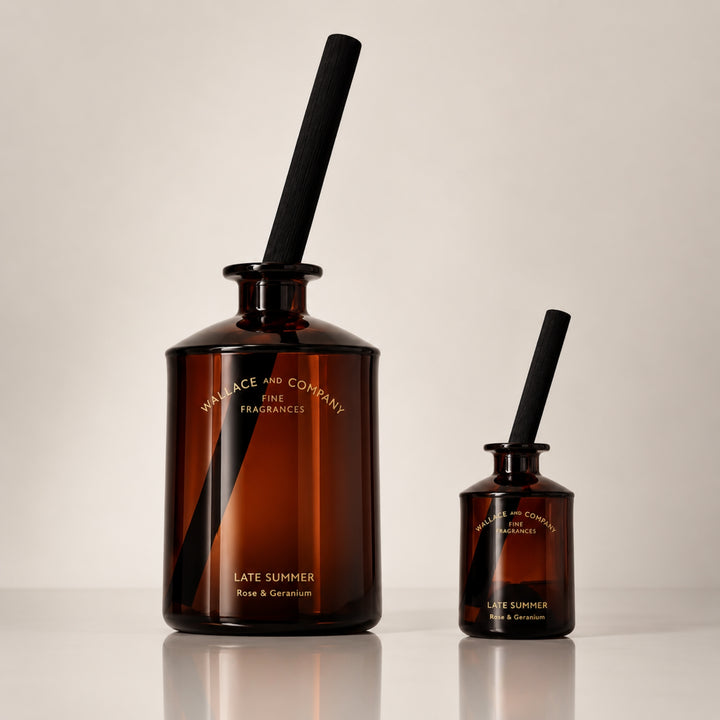 Large and small Wallace and Company “Late Summer – Rose & Geranium” reed diffusers in dark amber glass bottles with black diffuser sticks, displayed side by side against a warm neutral studio background.