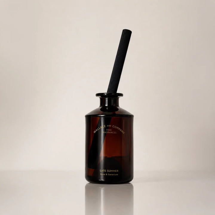 Wallace and Company “Late Summer – Rose & Geranium” reed diffuser in a dark amber glass bottle with a single black diffuser stick, photographed against a warm neutral studio background.