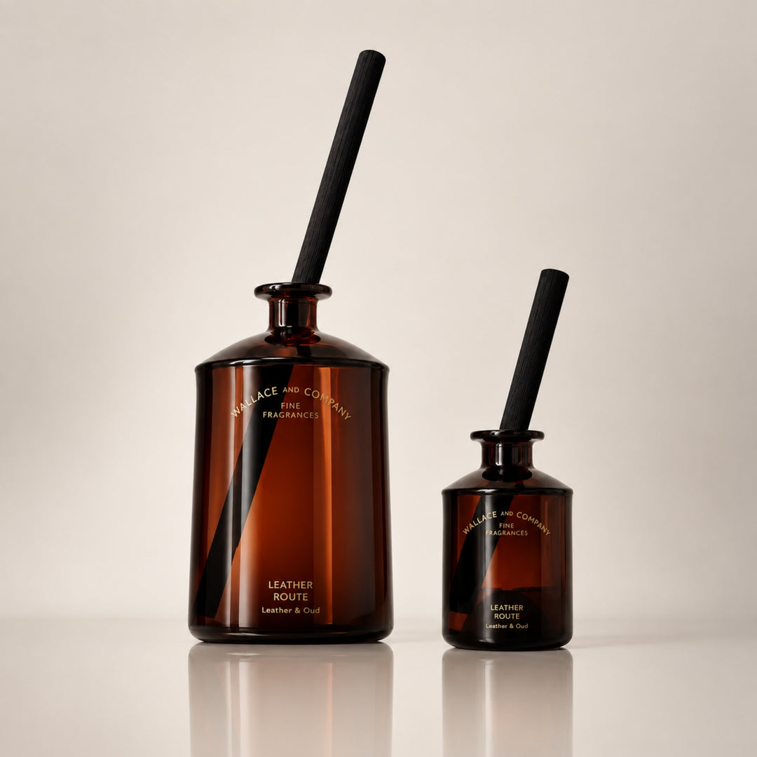 Large and small Wallace and Company “Leather Route – Leather & Oud” reed diffusers in dark amber glass bottles with black diffuser sticks, displayed side by side against a warm neutral studio background.