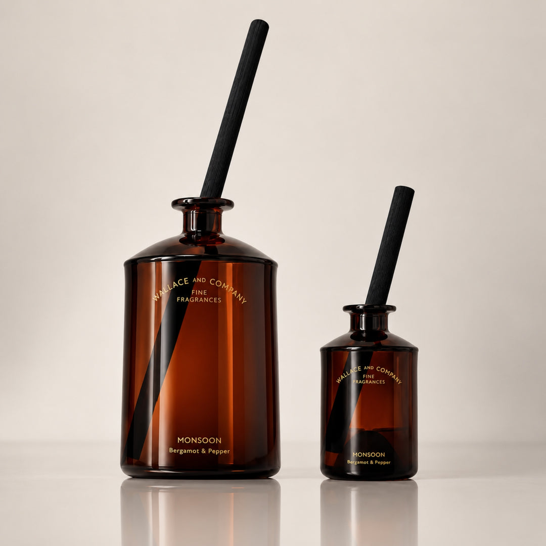 Large and small Wallace and Company “Monsoon – Bergamot & Pepper” amber-brown reed diffuser bottles displayed side by side with black diffuser sticks against a warm neutral studio background.