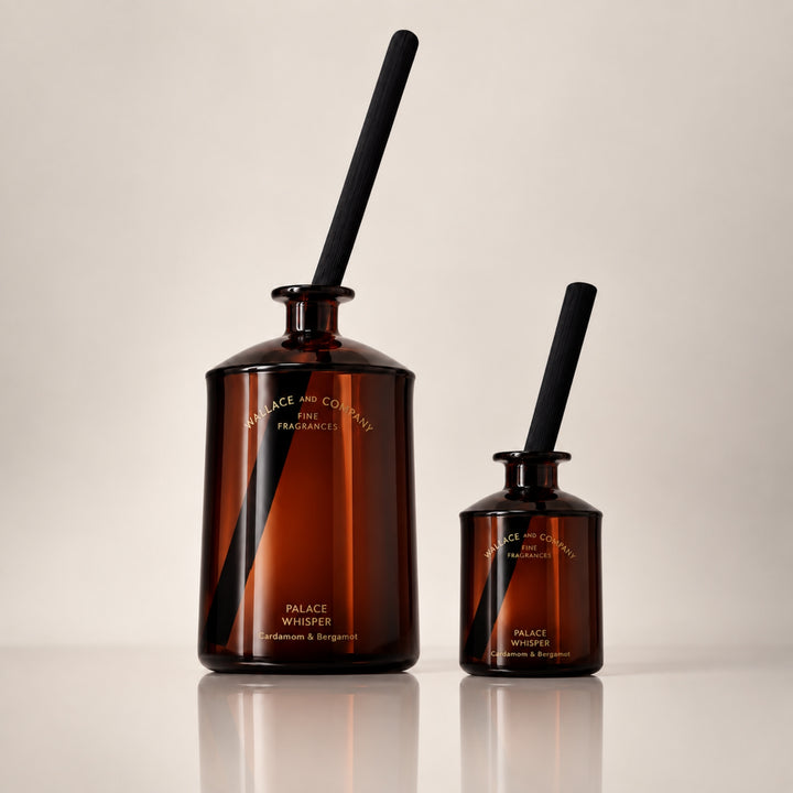 Large and small Wallace and Company “Palace Whisper – Cardamom & Bergamot” reed diffusers in dark amber glass bottles with black diffuser sticks, displayed side by side against a warm neutral studio background.