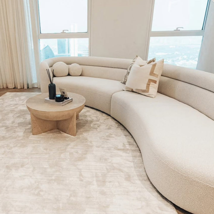 Bright, elegant living room featuring a large curved cream bouclé sofa with neutral cushions, a round natural wood coffee table, a soft beige area rug, and floor-to-ceiling windows. A black reed diffuser sits on the coffee table, adding a minimalist decorative accent.