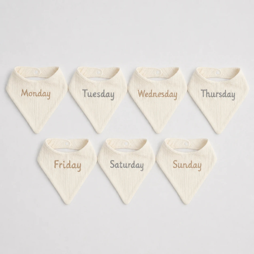 Set of seven cream-colored bibs with days of the week printed on them against a light gray background.