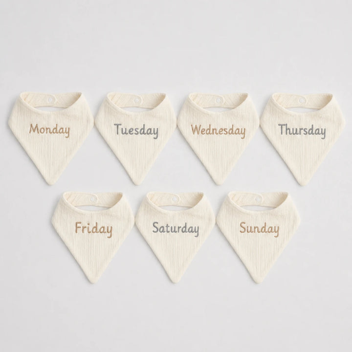 Set of seven cream-colored bibs with days of the week printed on them against a light gray background.