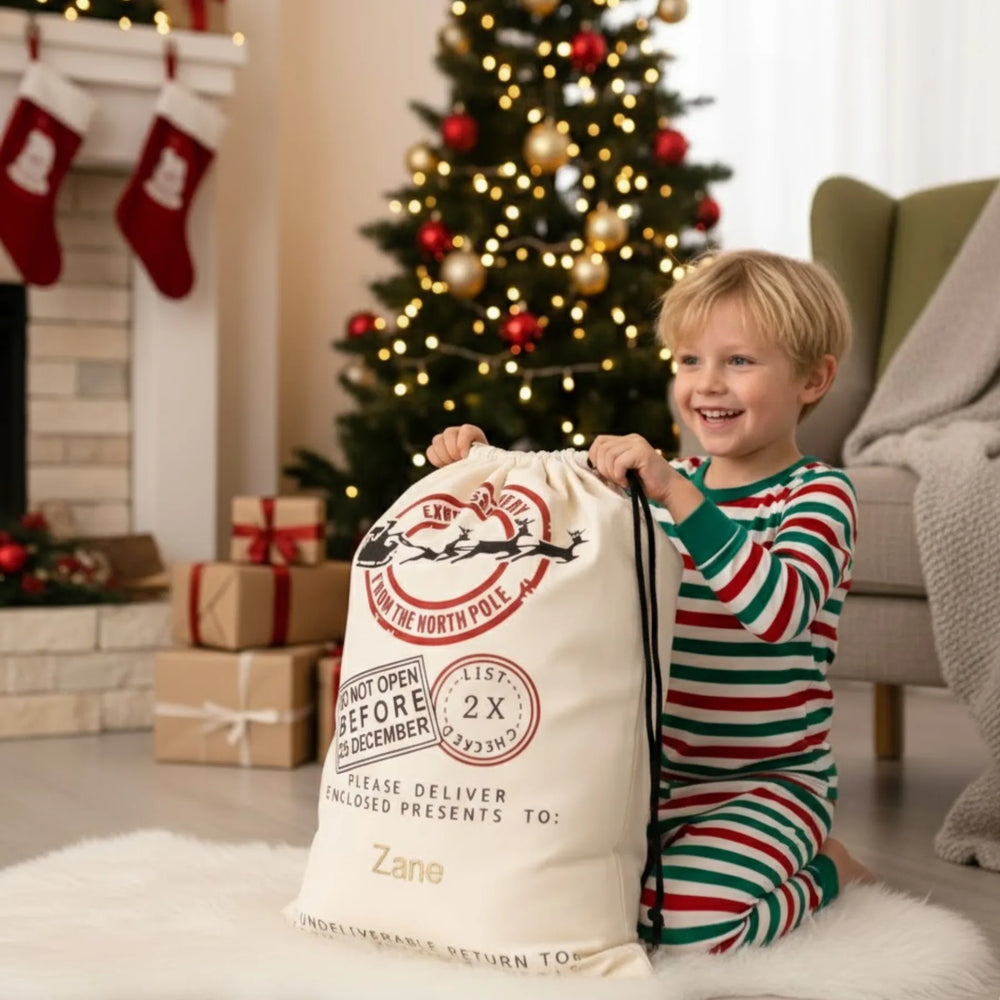 Child in pajamas holding a large Christmas present sack in a festive living room with a tree and stockings.
